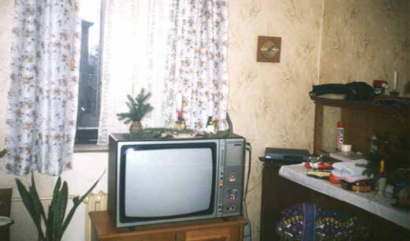 Blick in ein Zimmer, man sieht einen alten DDR-Fernsehen, ein Fenster mit Gardinen und eine Anrichte