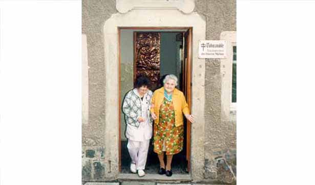 Eine Mitarbeiterin stützt eine ältere Bewohnerin beim Gang aus dem Haus