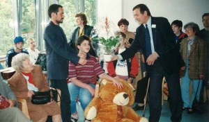 ein Mann übergibt einem Pfleger eine Blume, ein junger Bewohner sitzt dazwischen, er hält einen Plüschtierbären in der Hand, im gesamten Raum sind viele Menschen versammelt