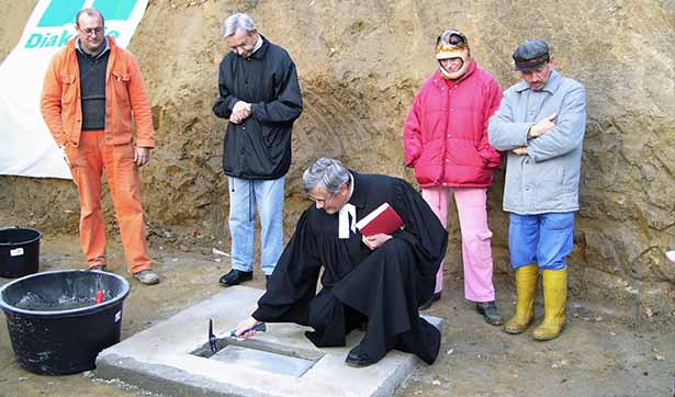Pfarrer Wachsmuth lägt den Grundstein, er klopft mit einem Hammer auf die Platte, daneben stehen drei Bewohner und der Bauleiter