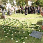 Mitarbeiter legen viele weiße Rosen auf den grünen Rasen, sie verbinden das Denkmal und die neue Gedenktafel, im Hintergrund stehen viele Mitarbeiter