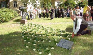Mitarbeiter legen viele weiße Rosen auf den grünen Rasen, sie verbinden das Denkmal und die neue Gedenktafel, im Hintergrund stehen viele Mitarbeiter