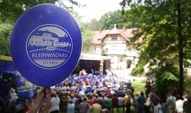 ein blauer Ballon mit der Aufschrift 125 Jahre Kleinwachau, im Hintergrund das Brunnenhaus und eine Bühne mit vielen Menschen davor sitzend