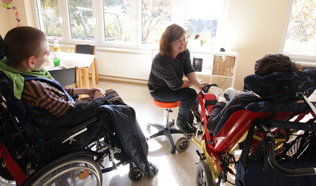 eine Betreuerin kümmert sich um zwei behinderte Menschen in speziellen Rollstühlen